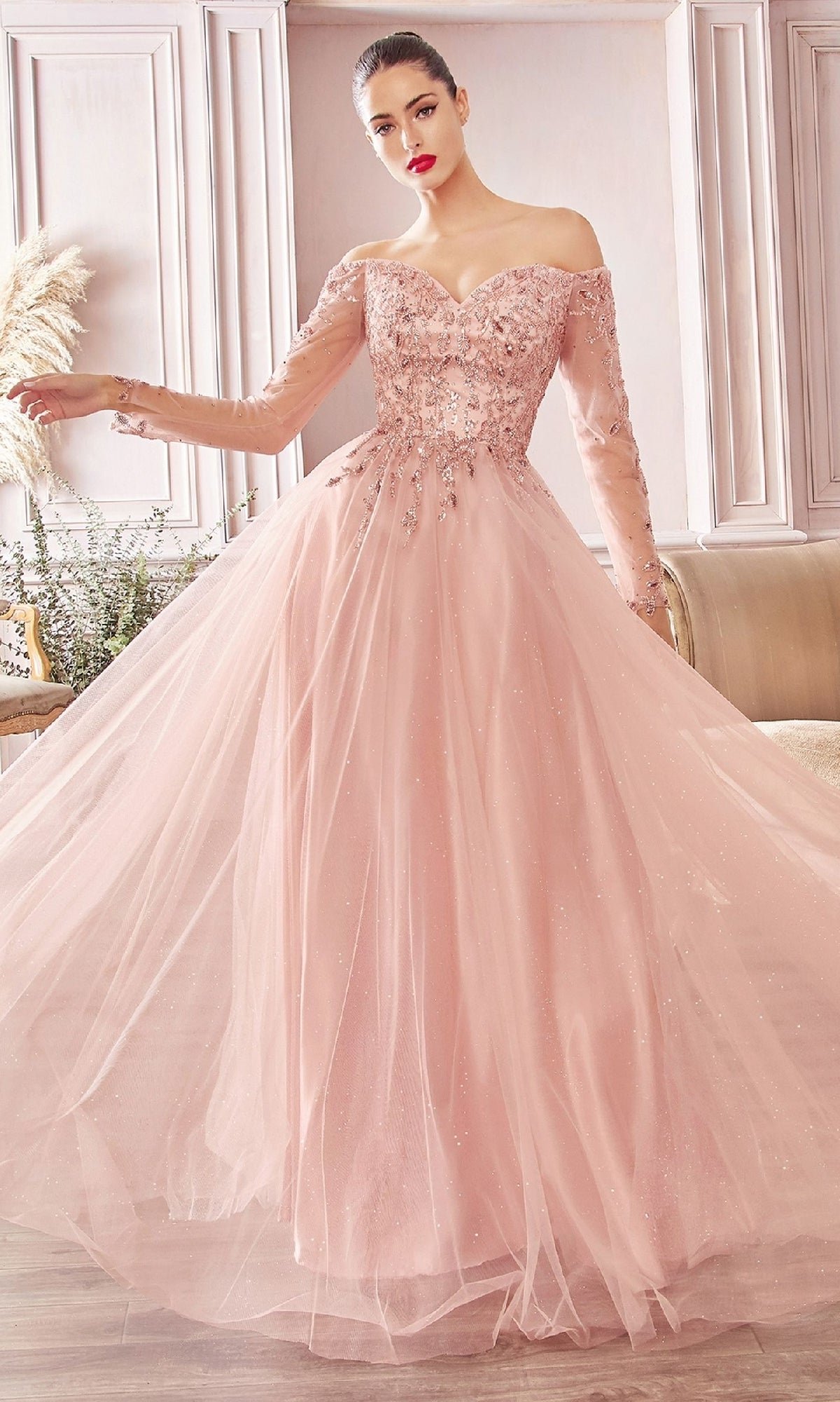 A woman stands indoors wearing the Ladivine CD0172, a blush pink long prom dress with sheer long sleeves, floral embellishments, and a full tulle skirt.